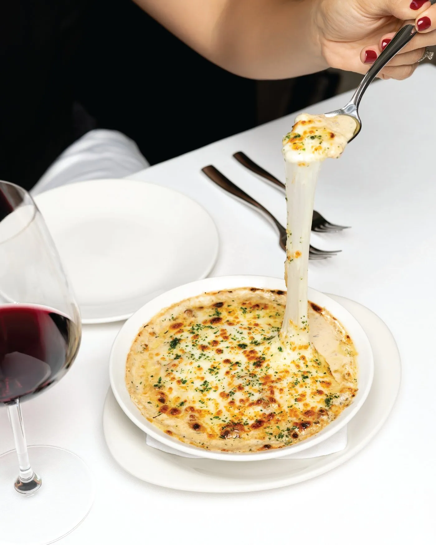 Una cuchara levanta anacates gratinados con queso fundido junto a una copa de vino tinto