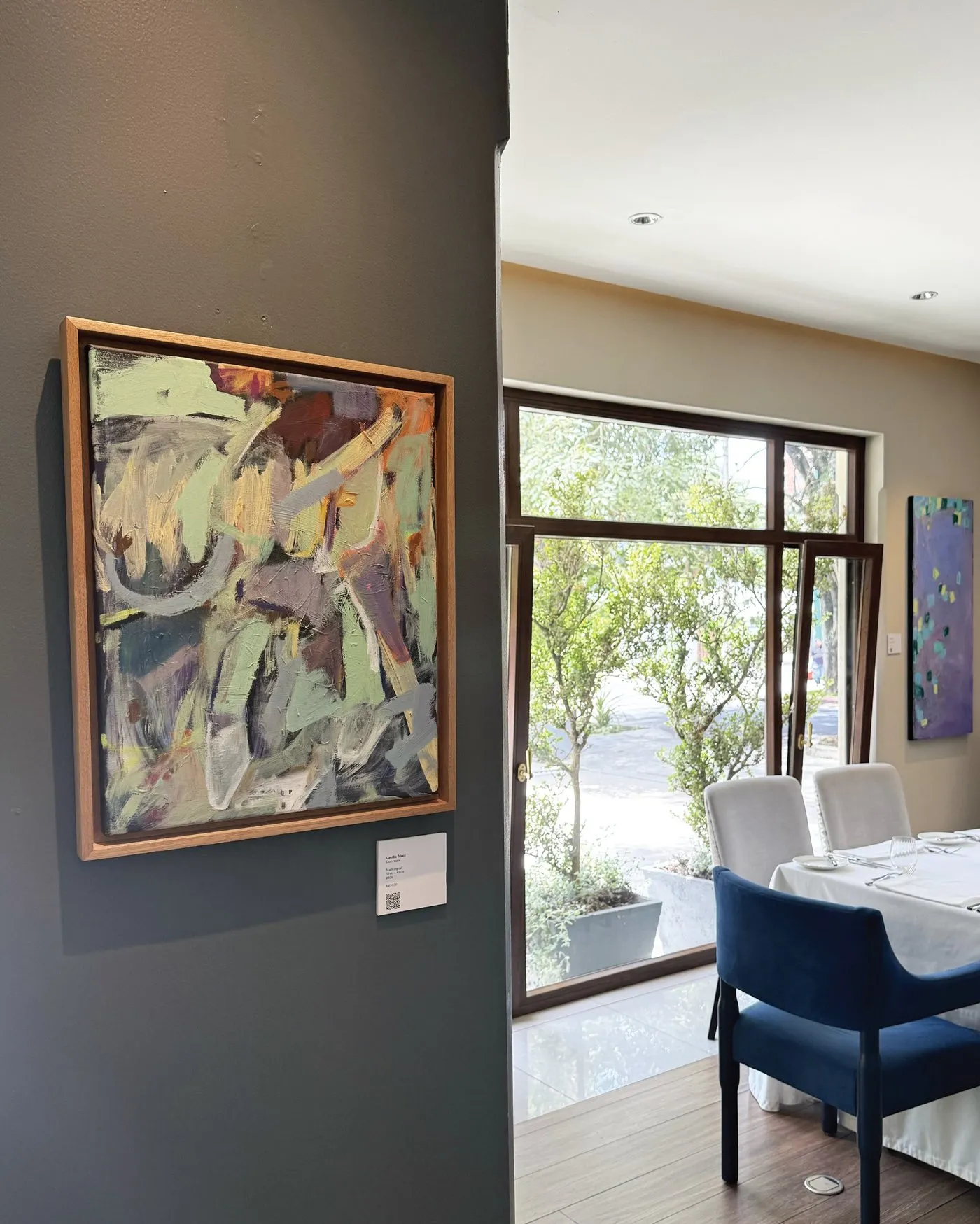 Pintura abstracta junto al ventanal, con mesas de mantel blanco y sillas azules al fondo