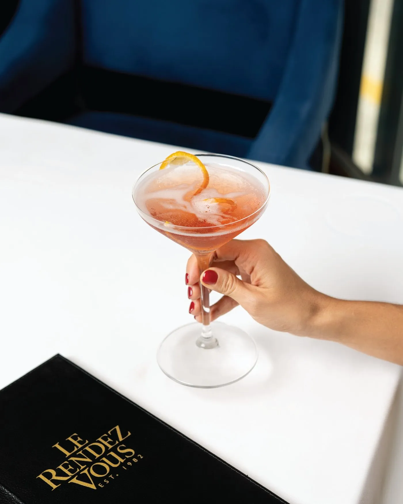 Una mano sostiene un cosmopolitan junto a la carta negra de Le Rendez Vous