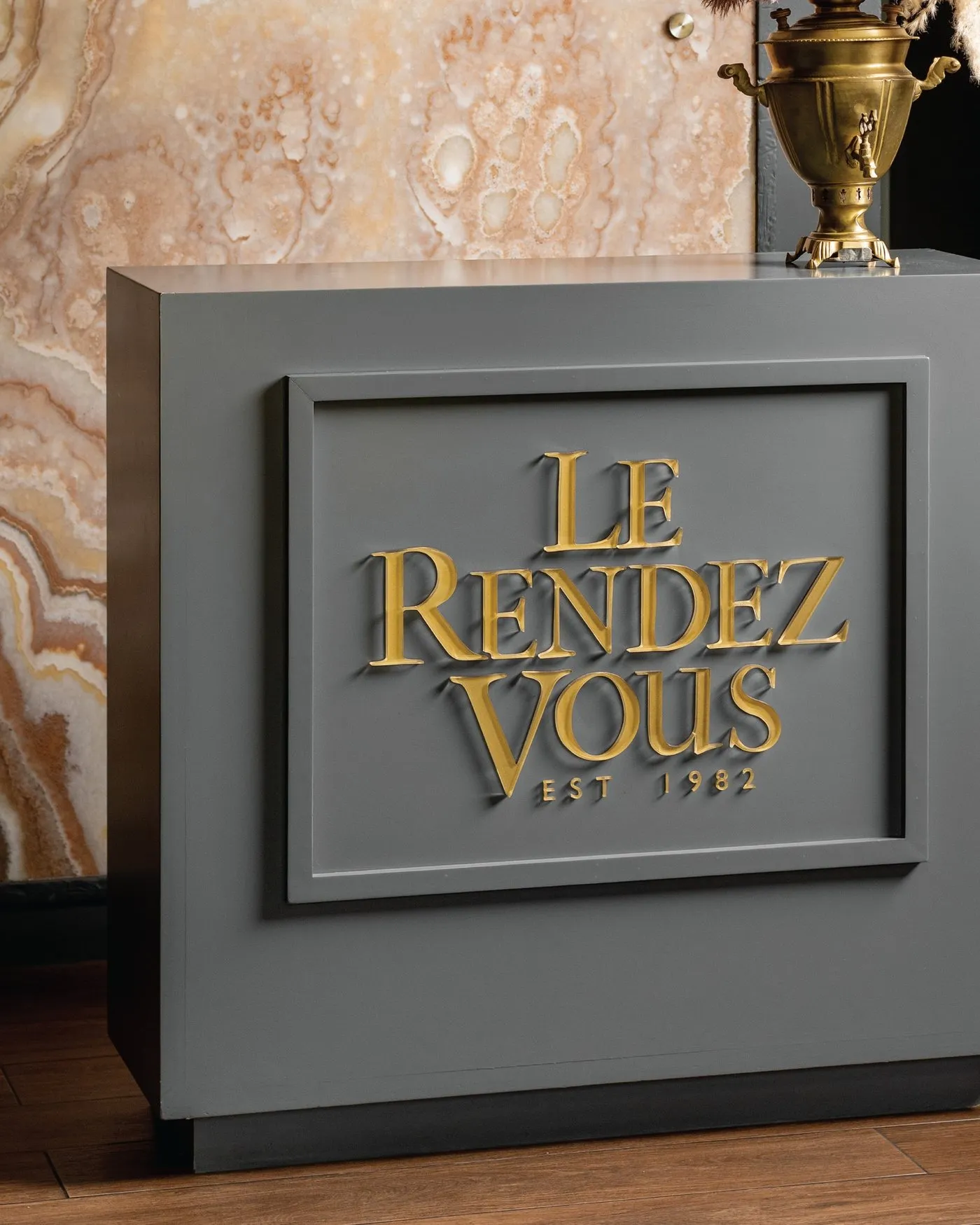 Letrero dorado de Le Rendez Vous, Est. 1982, sobre muro gris en la recepción